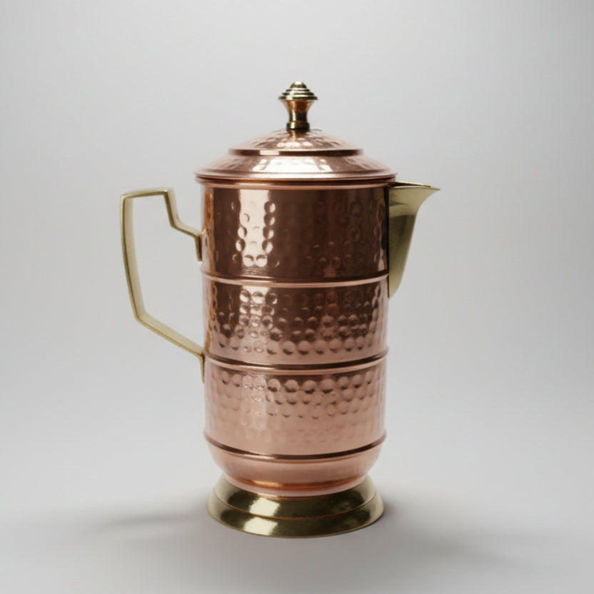 A copper jug with a hammer-textured surface, featuring a spout, handle, and a lid with a decorative knob on top.