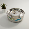 A small silver steel masala box with a lid, placed on a white surface, with a small plant pot and a toy visible in the background.