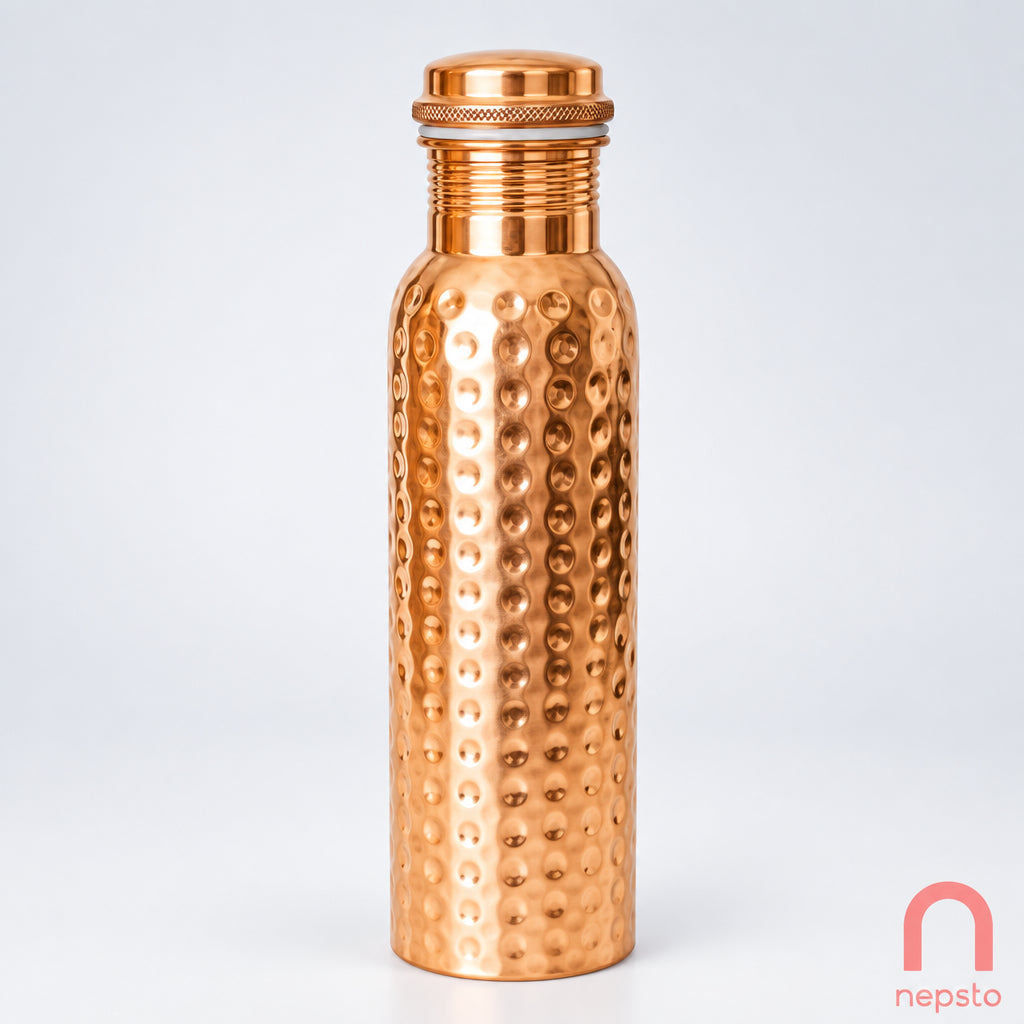 A copper bottle with a textured surface and a screw-on lid, placed on a white surface with a plant in the background.