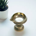 A brass diyo, traditional oil lamp, placed on a white surface with a plant pot in the background.