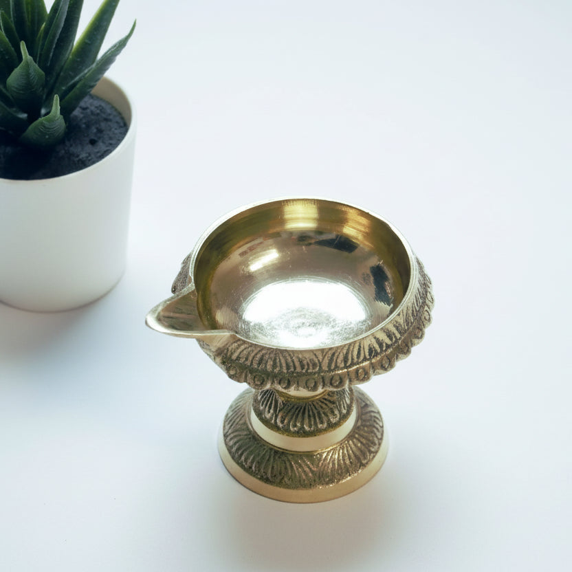 A brass diyo, traditional oil lamp, placed on a white surface with a plant pot in the background.