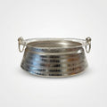 A hammered silver metal bowl with two side loop handles, resting on a white surface with a blurred grocery store background.