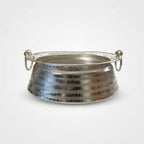 A hammered silver metal bowl with two side loop handles, resting on a white surface with a blurred grocery store background.