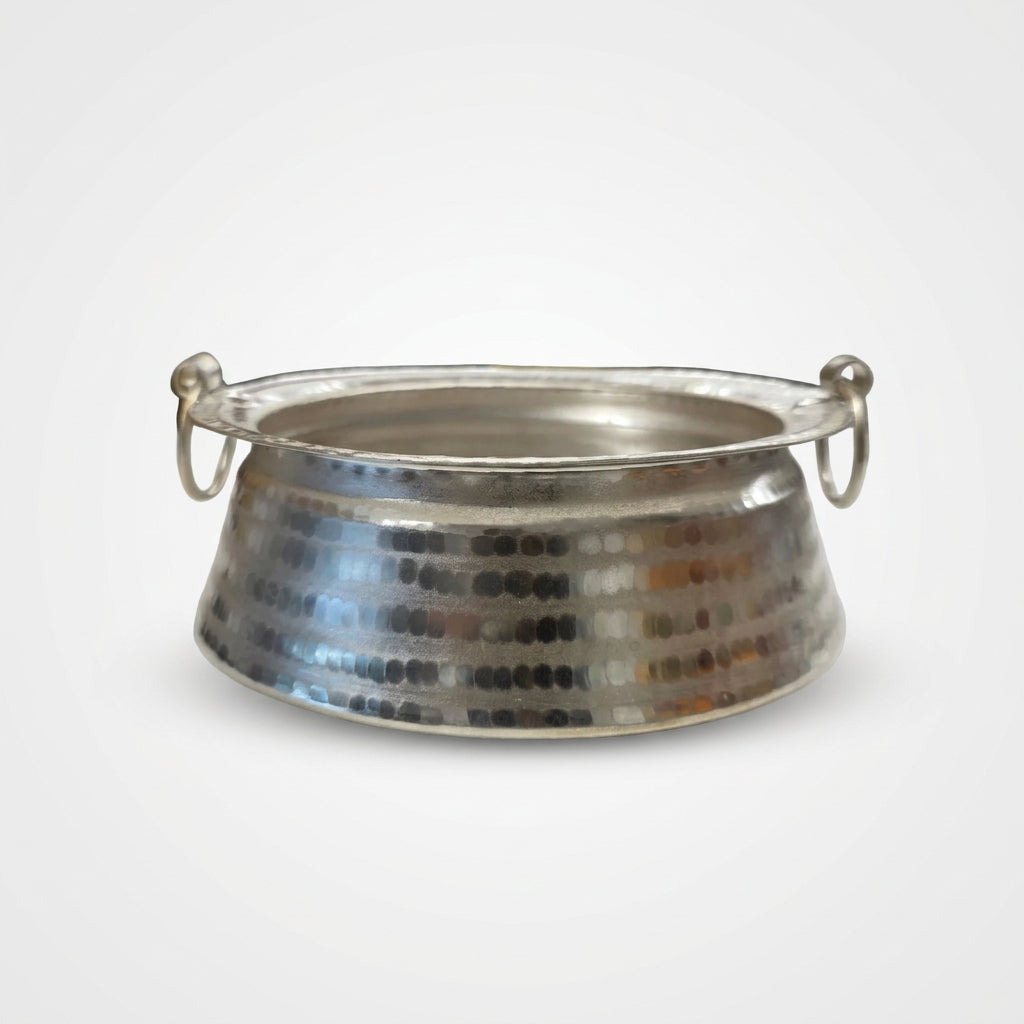 A hammered silver metal bowl with two side loop handles, resting on a white surface with a blurred grocery store background.