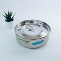 A large silver steel masala box with a lid, placed on a white surface, with a small potted plant in the background.